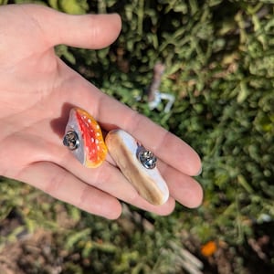 May include: Two handmade, colourful pins. One pin features a red and orange design with a silver pin back. The other pin has a white and brown design with a silver pin back. The pins are held in a hand.
