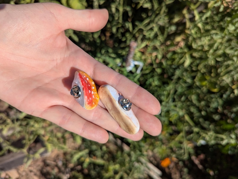 May include: Two handmade, colourful pins. One pin features a red and orange design with a silver pin back. The other pin has a white and brown design with a silver pin back. The pins are held in a hand.