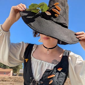 May include: A black witch's hat decorated with artificial mushrooms and green leaves. The hat is worn with a black choker necklace with a crystal pendant. The outfit includes a white blouse and a black vest with mushroom-shaped pins.