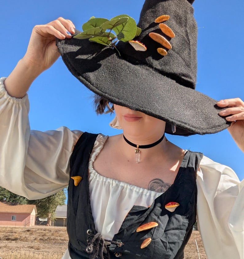 May include: A black witch's hat decorated with artificial mushrooms and green leaves. The hat is worn with a black choker necklace with a crystal pendant. The outfit includes a white blouse and a black vest with mushroom-shaped pins.