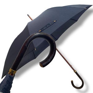 May include: A navy blue umbrella with a curved, dark wooden handle and a matching wooden shaft. The umbrella's canopy has a textured pattern and is accented with gold-coloured hardware. The handle is approximately 15 cm wide.