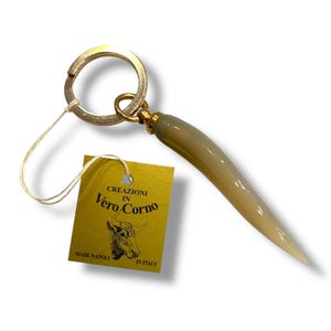 May include: A gold-coloured keyring with a horn-shaped pendant. The pendant is a gradient of light beige to white. A yellow tag reads "CREAZIONI IN Vero Corno MADE NAPOLI IN ITALY" with a bull illustration.