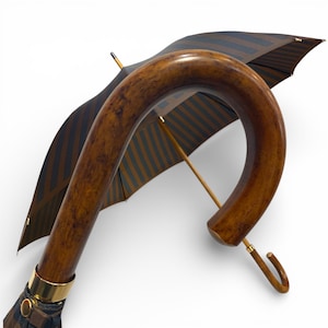 May include: A traditional umbrella featuring a curved wooden handle and a striped canopy in brown and blue. The handle has a varnished finish, and a gold-coloured metal band accents the base. The umbrella's design suggests a focus on traditional style and craftsmanship.