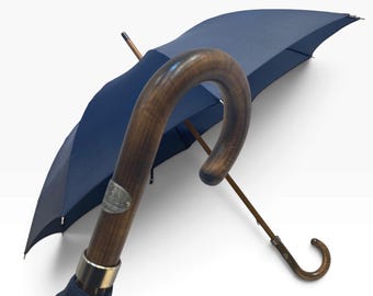 Puede incluir: Un paraguas azul marino con un mango de madera curvado y un emblema de metal plateado. El paraguas tiene un eje de madera y un segundo mango de madera más pequeño unido a la estructura, ofreciendo un diseño de doble mango.