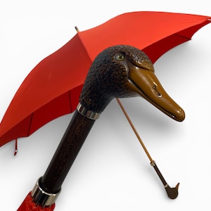May include: A red umbrella featuring a duck head handle. The handle is dark brown with a silver band. The duck head is brown and tan with a textured finish. The umbrella is open, displaying its bright red canopy.