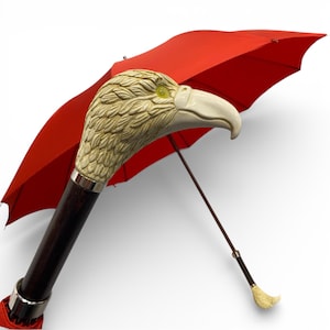 May include: A red umbrella featuring a decorative eagle head handle. The handle is dark brown with a silver band. The eagle head is beige with a white beak and yellow eye. The umbrella is open.