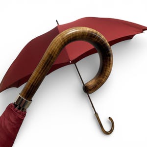 May include: A burgundy umbrella with a curved wooden handle. The handle features a dark wood grain pattern. The umbrella's canopy is a solid burgundy colour, and the metal accents are silver.