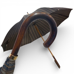 May include: A traditional umbrella with a curved wooden handle and a striped canopy in brown and beige. The handle has a gold-coloured metal band. The umbrella is partially open, displaying its design.