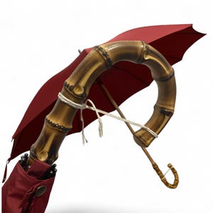 May include: A red umbrella featuring a curved bamboo handle. The handle is light brown with darker brown detailing. The umbrella is open, displaying the taut red fabric. White string secures the handle.