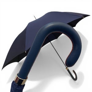 May include: A navy blue umbrella with a curved handle. The handle is wrapped in blue leather with red stitching. A gold-coloured metal band accents the base of the handle. The umbrella's canopy is a deep navy colour.