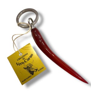 May include: A red horn-shaped keyring with a silver ring and a yellow tag. The tag reads "CREAZIONI IN Vero Corno" with a bull illustration and "MADE NAPOLI IN ITALY". The horn has a smooth, glossy finish.