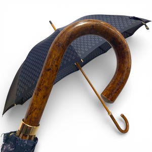 May include: A partially open umbrella with a dark blue canopy and a white floral pattern. The curved handle is varnished wood, and the shaft is lighter wood. A gold-coloured metal band accents the base.