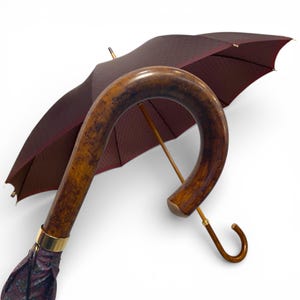 May include: An elegant umbrella featuring a curved wooden handle and a burgundy canopy. The canopy has a subtle pattern and is trimmed with gold-coloured accents. The handle is a rich, warm brown, and the umbrella is open, displaying its sophisticated design.