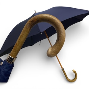 May include: A navy blue umbrella with a curved wooden handle. The handle has a textured, light brown finish. The umbrella's canopy is open, revealing its dark blue fabric. A silver metal band secures the handle to the fabric.