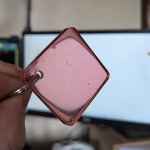 May include: A pink square keychain with a hole for a ring. The keychain is made of clear plastic with a slight pink tint.