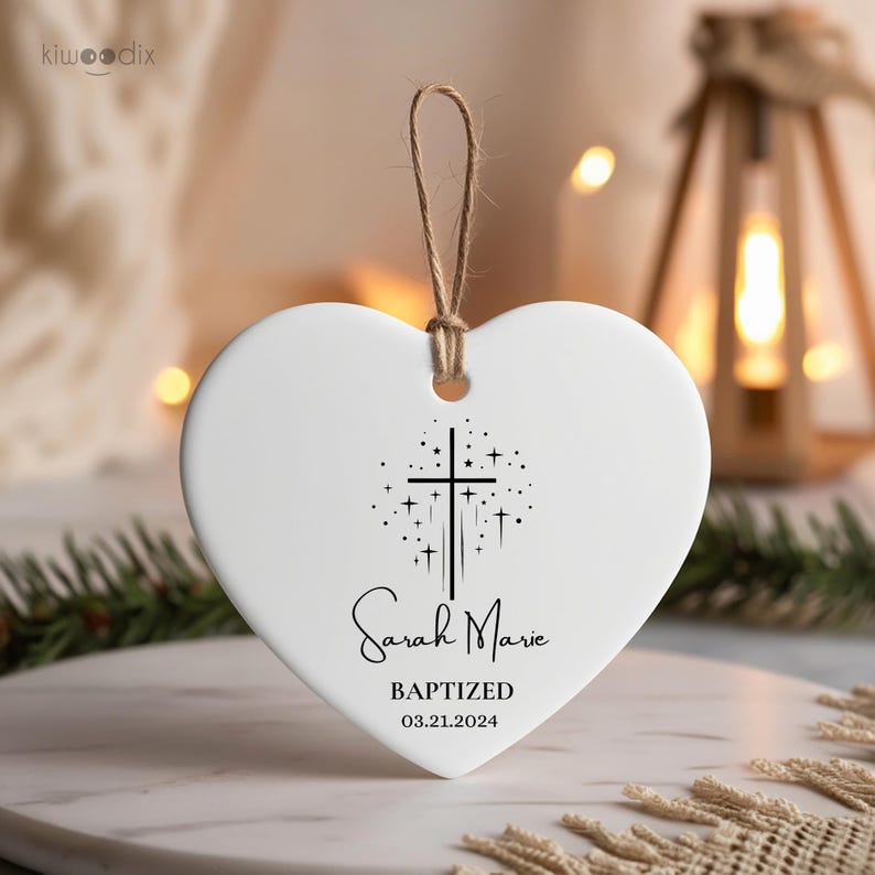 May include: White heart-shaped ceramic ornament with a black cross and stars design. The ornament reads "Sarah Marie Baptized 03.21.2024".