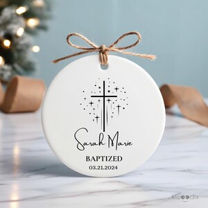 May include: White ceramic ornament with a black cross and stars design. The text "Sarah Marie Baptized 03.21.2024" is printed on the ornament.