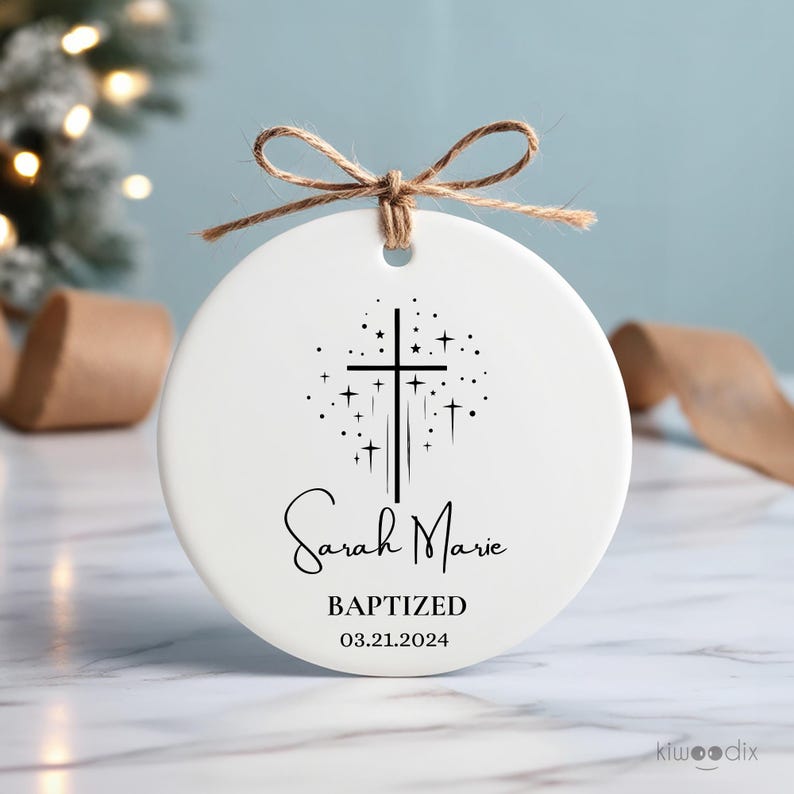 May include: White ceramic ornament with a black cross and stars design. The text "Sarah Marie Baptized 03.21.2024" is printed on the ornament.