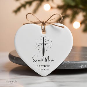May include: White ceramic heart-shaped ornament with a black cross and stars design. The ornament reads "Sarah Marie Baptized 03.21.2024".