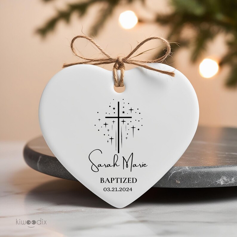 May include: White ceramic heart-shaped ornament with a black cross and stars design. The ornament reads "Sarah Marie Baptized 03.21.2024".