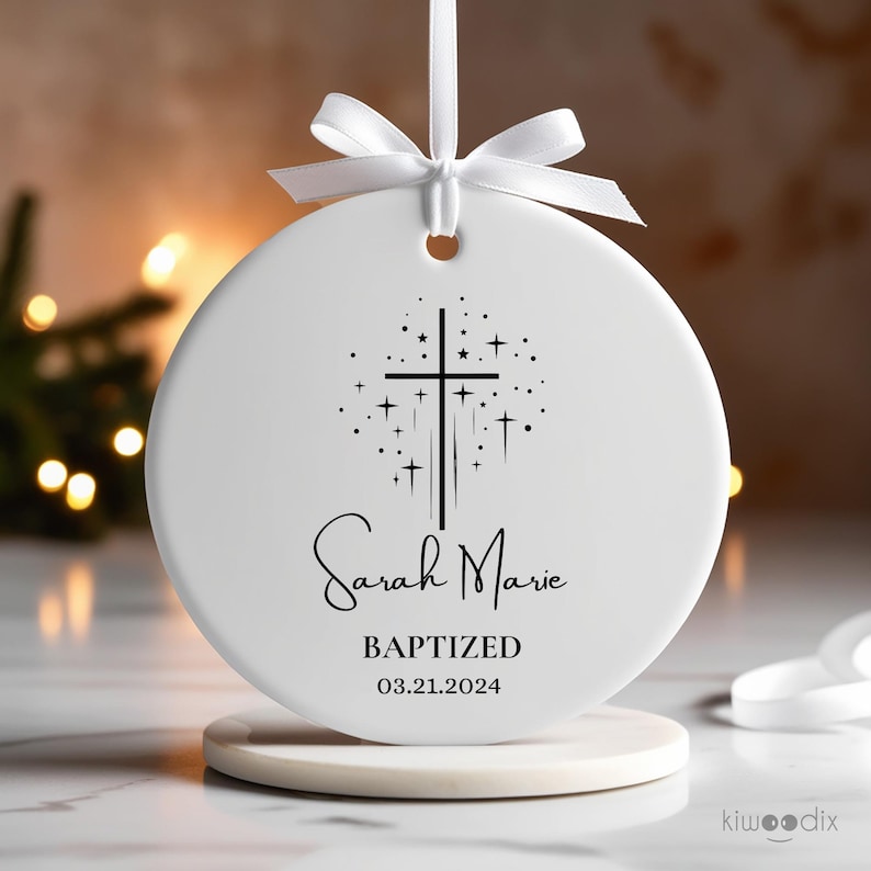 May include: White ceramic ornament with a black cross and stars design. The text "Sarah Marie Baptized 03.21.2024" is printed on the ornament.