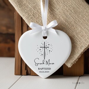 May include: White ceramic heart-shaped ornament with a black cross and stars design. The ornament is personalized with the name "Sarah Marie" and the text "Baptized 03.21.2024".