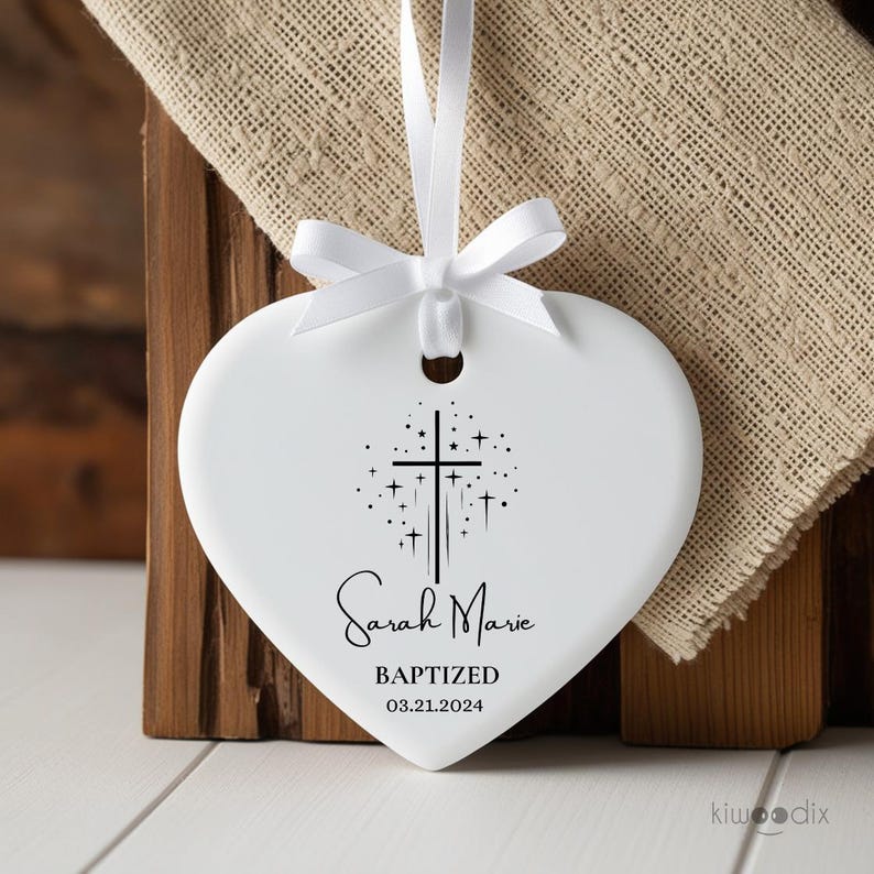 May include: White ceramic heart-shaped ornament with a black cross and stars design. The ornament is personalized with the name "Sarah Marie" and the text "Baptized 03.21.2024".