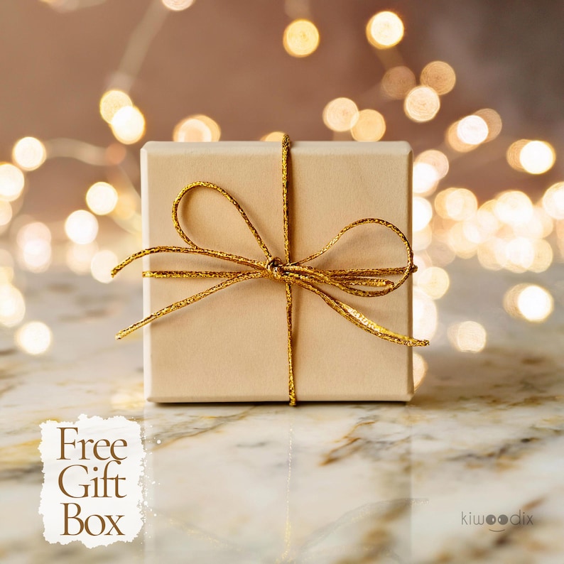 May include: A small, beige gift box tied with a gold ribbon and bow. The box is sitting on a white marble surface with a blurred background of golden lights.