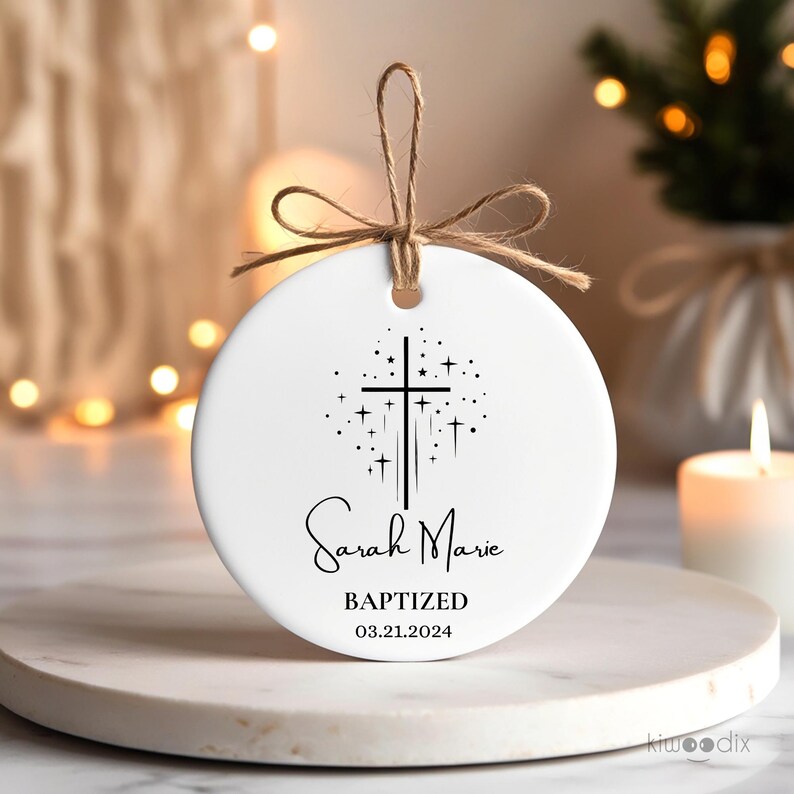 May include: White ceramic ornament with a black cross and stars design. The text "Sarah Marie Baptized 03.21.2024" is printed on the ornament.