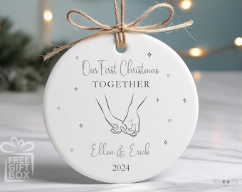 May include: White ceramic Christmas ornament with black text that reads "Our First Christmas Together" and "Ellen & Erick 2024". A simple line drawing of two hands holding each other is featured on the ornament.