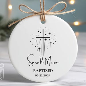 May include: White ceramic ornament with a black cross and stars design. The text "Sarah Marie Baptized 03.21.2024" is printed on the ornament.