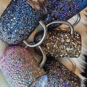 Może przedstawiać: Kolekcja zdobionych kubków podróżnych w różnych kolorach, w tym niebieskim, fioletowym, złotym, różowym i zielonym. Każdy kubek jest pokryty błyszczącymi kryształkami i posiada srebrny uchwyt oraz przezroczystą pokrywkę.