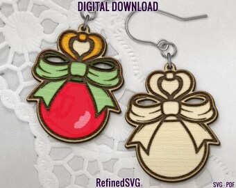 May include: Wooden Christmas ornament earrings. One earring is painted red with a green bow and gold accents. The other earring is unpainted. The earrings are hanging from silver hooks. The words "DIGITAL DOWNLOAD" and "RefinedSVG" are visible.