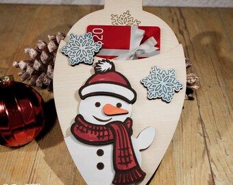 Può includere: Addobbo natalizio in legno a forma di lampadina con un design a pupazzo di neve. L'addobbo ha una tasca per una carta regalo e presenta fiocchi di neve e il testo "20 €".