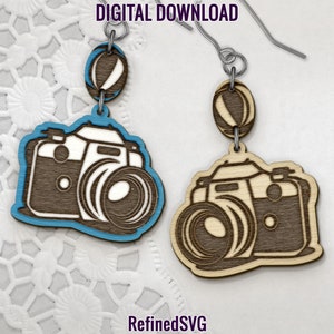 Puede incluir: Pendientes de madera con diseño de cámara. Un par tiene un contorno azul, el otro es de madera natural. Los pendientes cuelgan de ganchos plateados. Se ven las palabras "DIGITAL DOWNLOAD" y "RefinedSVG".