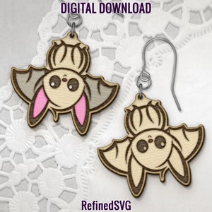 May include: A pair of wooden bat earrings with a cute cartoon bat design. The bats are brown with gray wings and pink ears. The earrings are hanging from silver hooks.