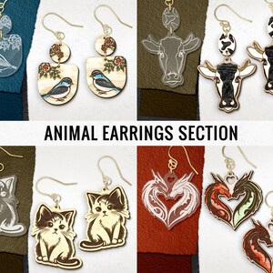 May include: A collection of animal-themed earrings. Designs include birds, cows, cats, and dragons. The earrings are made from wood and acrylic, with gold-colored ear wires. The text "ANIMAL EARRINGS SECTION" is displayed below the earrings.