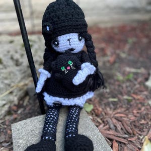 May include: A handmade crochet doll with a black dress, hat, and braids. The doll has pale skin, a stitched mouth, and is holding a black cat with green and pink accents. The doll is sitting on a concrete block.