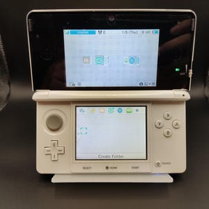 May include: A white handheld gaming console with a dual-screen design. The top screen displays a digital interface with icons, while the bottom touchscreen shows a menu with the text Create Folder. It features a directional pad, buttons, and a joystick.