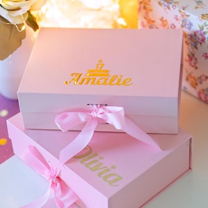 May include: Two rectangular light pink gift boxes with hinged lids and matching pink ribbon closures. The top box features the gold foil name Amalie with a birthday cake icon, while the bottom box displays the gold glitter name Olivia.