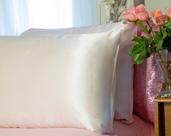 May include: Two white satin pillowcases on a pink bedspread. The pillowcases are smooth and wrinkle-free. A vase of pink roses is on a table in the background.