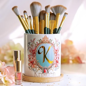 May include: A collection of makeup brushes with gold handles and white bristles in a decorative ceramic holder. The holder features a floral design, a gold letter "K", and the name "Kristen". A small bottle of makeup is in front.