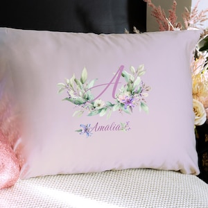 May include: A light pink pillow with a floral design and the letter "A" in a lavender color. The pillow has the name "Amalia" written in a script font below the floral design. The pillow is on a white surface.