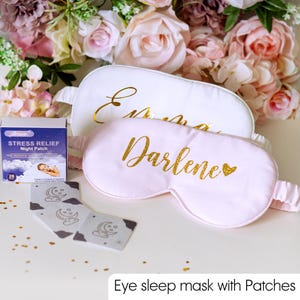 May include: White and pink eye sleep masks with gold lettering, one reading "Emma" and the other "Darlene". A box of stress relief night patches and a set of eye patches are also present. The background features a floral arrangement.