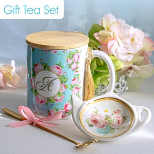 May include: A gift tea set featuring a floral-decorated mug with a wooden lid, a gold spoon with a pink bow, and a teapot-shaped tea bag holder. The mug has the letter "A" in a floral wreath. The set is on a white surface.