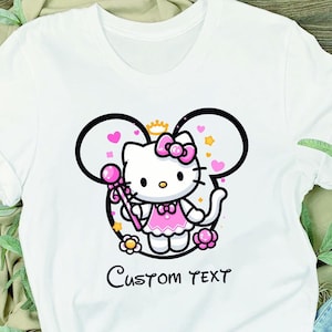 May include: White t-shirt with a pink Hello Kitty character wearing a pink dress and holding a wand. The character is inside a black outline of Mickey Mouse ears with pink hearts and flowers. The text "Custom Text" is printed below the character.