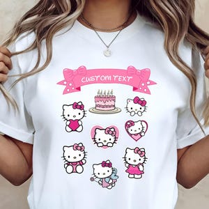May include: White t-shirt featuring multiple Hello Kitty designs in pink and red. A pink banner with the text "CUSTOM TEXT" is above a cake with lit candles. The shirt has a crew neck and short sleeves.