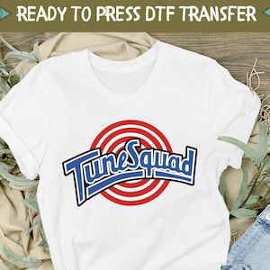 May include: White t-shirt featuring a blue and red "Tune Squad" graphic with the iconic "What's Up Doc?" logo. The shirt is displayed on a wooden surface with a pair of white trainers and blue jeans.