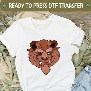 May include: White t-shirt with a cartoon illustration of a beast's face in brown tones. The text "READY TO PRESS DTF TRANSFER" is at the top. The shirt is styled with blue jeans and white trainers, set on a wooden surface.