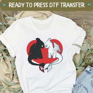 May include: White t-shirt featuring a black and white dragon design inside a red heart. The shirt has a crew neck and short sleeves. The text "READY TO PRESS DTF TRANSFER" is at the top. The shirt is paired with blue jeans and white sneakers.